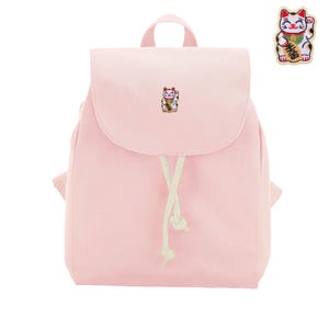 Może przedstawiać: Jasnoróżowy plecak z małą haftowaną naszywką kota maneki-neko na przedniej klapie. Torba posiada górny uchwyt, zapięcie na sznurek z białymi wiązaniami oraz pasującą naszywkę kota maneki-neko widoczną w prawym górnym rogu obrazu.