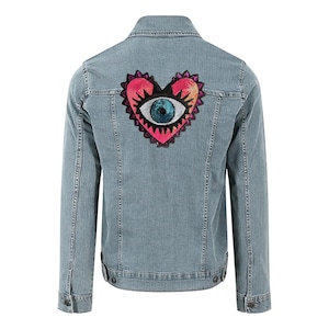 Op de afbeelding: Lichtblauwe spijkerjas met een hartvormige patch op de rug. De patch heeft een oogontwerp met roze, oranje en gele accenten en een blauw paillettoog.