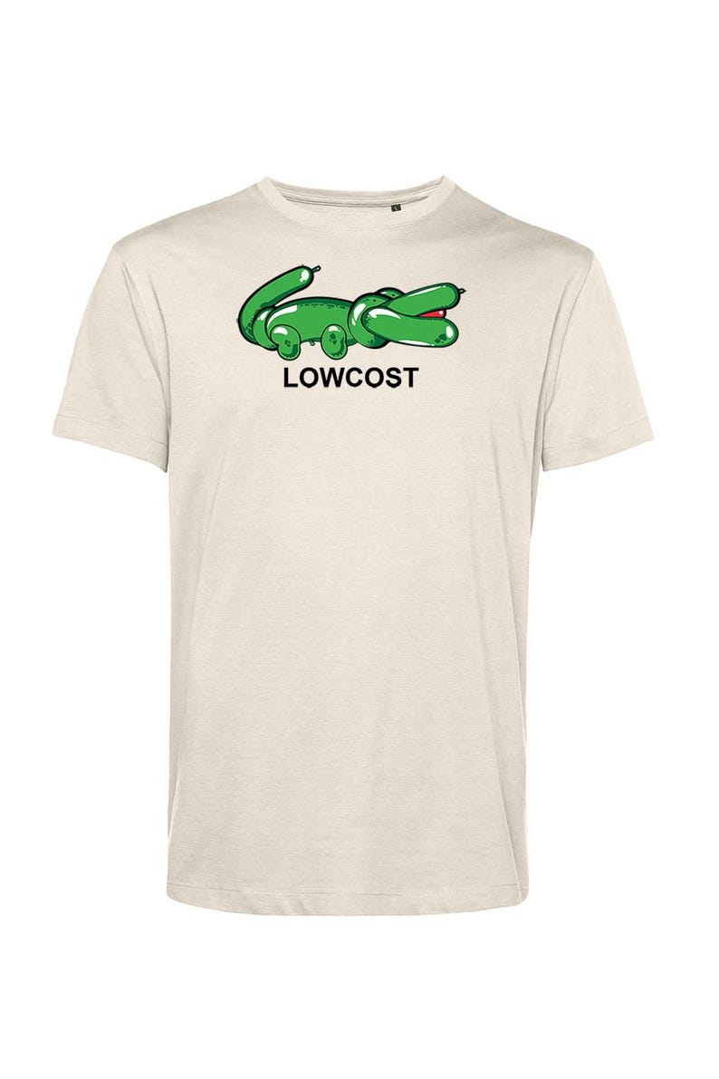 May include: A light beige t-shirt featuring a green cartoon alligator design with the word "LOWCOST" in black text. The alligator has a red mouth and is positioned on the front of the shirt. The shirt has short sleeves.