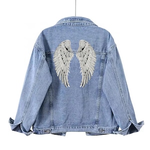 Puede incluir: Chaqueta vaquera azul claro con alas de ángel de lentejuelas plateadas en la espalda. La chaqueta tiene cuello clásico, cierre de botones en la parte delantera y mangas largas. Las alas están detalladas con lentejuelas individuales.