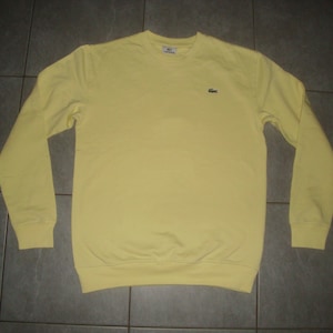 Op de afbeelding: Een lichtgeel sweatshirt met lange mouwen en een ronde hals. Het shirt heeft een klein groen krokodillenlogo op de borst. De manchetten en zoom zijn geribbeld. Het shirt is gemaakt van een zacht materiaal.