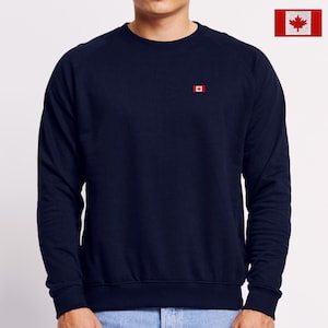 May include: A dark navy blue long-sleeve sweatshirt featuring a small embroidered Canadian flag on the left chest. The garment has a crew neckline, ribbed cuffs, and a ribbed hem. A small inset shows a larger view of the red and white maple leaf flag.