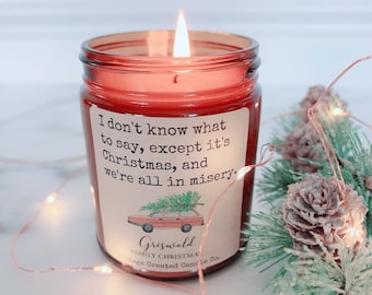 May include: A lit red candle in a glass jar with a label that reads "I don't know what to say, except it's Christmas, and we're all in misery." The label also features a car with a Christmas tree on top. The candle is surrounded by pine needles and fairy lights.