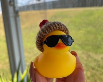 May include: A yellow rubber duck wearing a brown knitted hat with a red pom-pom and black sunglasses.