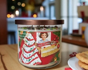May include: A glass candle jar with a copper-coloured lid. The label features a vintage illustration of a cake and a girl, with the text "Little Debbie Christmas Tree Cakes". The candle is on a wooden surface.