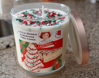 May include: A white candle in a clear glass jar with a lid. The candle is topped with red and green sprinkles. The jar has a label that says "Little Debbie Christmas Tree Cakes".