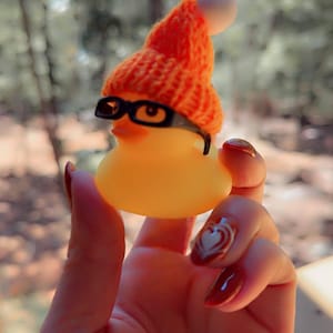May include: A yellow rubber duck wearing a small orange knitted hat with a white bobble and black-rimmed glasses. The duck is held in a hand with red nail varnish and a heart design.