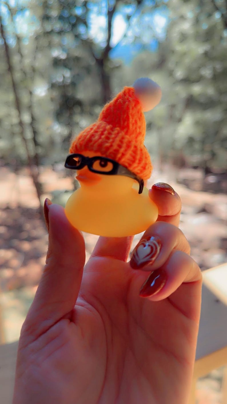 May include: A yellow rubber duck wearing a small orange knitted hat with a white bobble and black-rimmed glasses. The duck is held in a hand with red nail varnish and a heart design.
