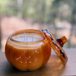 May include: A pumpkin-shaped candle in a glass jar with a lid. The candle is orange with three white wicks. The lid is open, revealing the candle inside. The jar sits on a wooden surface.