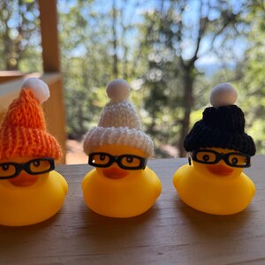 May include: Three yellow rubber ducks, each with black-rimmed glasses and a knitted hat. The hats are orange, white, and black, with a white bobble. The ducks are on a wooden surface.