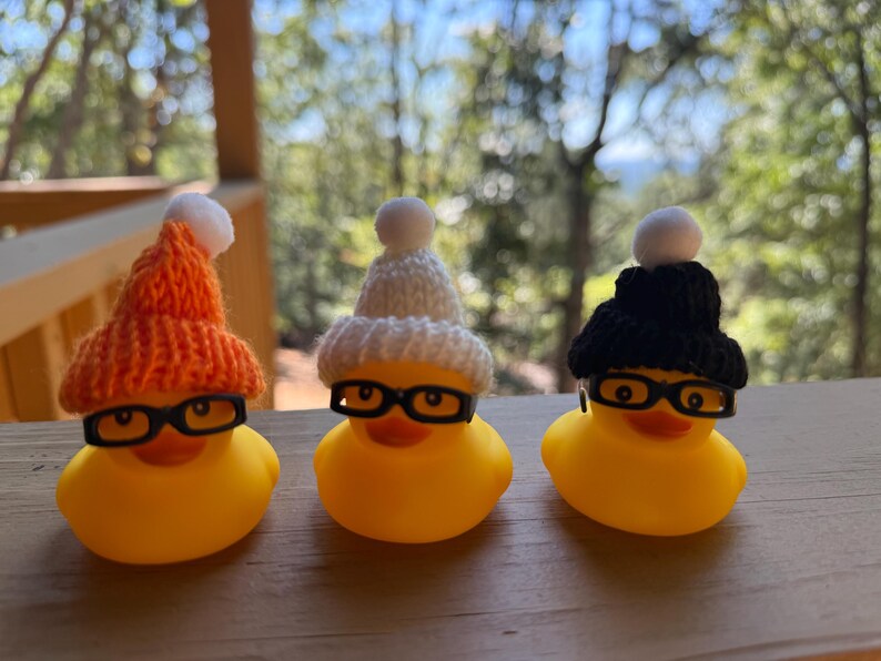 May include: Three yellow rubber ducks, each with black-rimmed glasses and a knitted hat. The hats are orange, white, and black, with a white bobble. The ducks are on a wooden surface.