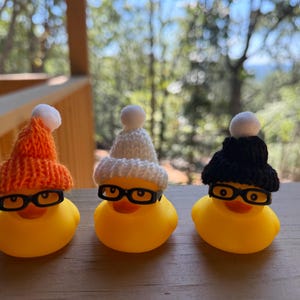 May include: Three yellow rubber ducks, each wearing black-rimmed spectacles and knitted hats in orange, white, and black, topped with white pom-poms. They are displayed on a wooden surface.