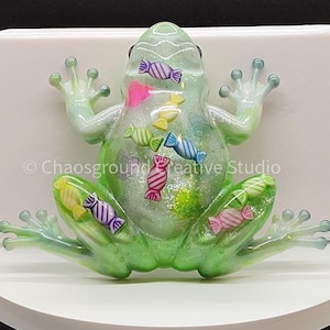 May include: A whimsical, green frog figurine with candy-shaped embellishments. The frog has a translucent body with colorful candies inside. The frog is sitting on a white surface.