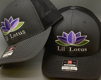May include: Two black and gray trucker hats with a purple and green embroidered lotus flower and the text "Lil Lotus". The hat on the right has a black and red label with the text "Richardson. Est. 1970 Original 112 Trucker Adjustable".