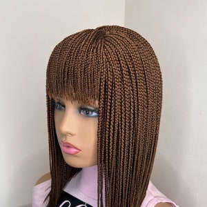 May include: A brown braided wig with a blunt fringe, displayed on a mannequin head. The wig features small, tightly woven braids and a natural-looking hairline. The mannequin is wearing a light pink shirt with the text "Ella's Beauty Lounge...your beauty...your pride".