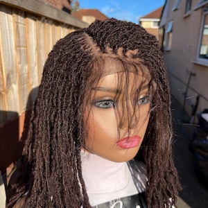 May include: A brown micro-braided wig displayed on a mannequin head. The wig has a realistic hairline and is styled with small, individual braids. The mannequin is wearing a white turtleneck. The background shows a wooden fence and a sunny sky.