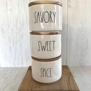 May include: A stack of three white ceramic canisters with wooden lids, labeled "SAVORY", "SWEET", and "SPICE" in black script. The canisters are stacked on a small, square wooden tray. The background is a light-colored wooden surface.