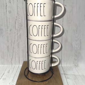 May include: A stack of four white ceramic coffee mugs with the word "COFFEE" in black lettering. The mugs are held in a black metal stand and sit on a wooden base. The background is a light wood-paneled wall.