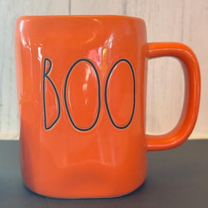 Puede incluir: Taza de cerámica naranja brillante con la palabra "BOO" en letras negras. La taza tiene un asa curva y un acabado brillante. Una taza con temática de Halloween.