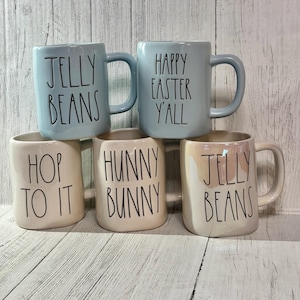 Puede incluir: Cinco tazas de cerámica con lemas de temática pascual. Las tazas son de dos colores: azul claro y beige claro con un esmalte moteado. Los lemas en las tazas son: "Jelly Beans", "Happy Easter Y'all", "Hop To It", "Hunny Bunny" y "Jelly Beans".