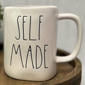 May include: Cream-colored ceramic mug with a curved handle. The mug features the words "SELF MADE" in black, handwritten-style lettering. The mug is sitting on a wooden tray.