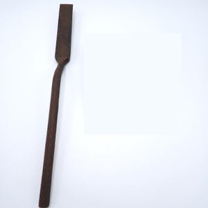 May include: A long, narrow, dark brown metal tool with a flat, rectangular head and a straight, slender handle. The surface of the metal appears weathered and textured, showing signs of age and oxidation throughout the entire length of the object.