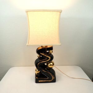 May include: A table lamp featuring a square, off-white fabric shade and a glossy black ceramic base with a zigzag, serpentine shape. The base is accented with polished gold-colored spheres and a gold-toned pull chain for the light switch.