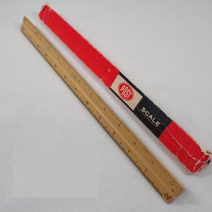 May include: A tan triangular architect scale ruler with black numerical markings and a corresponding long rectangular red, white, and black cardboard storage box. The box features the brand name POST and the word SCALE printed in white text.