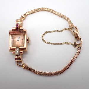 May include: A gold-tone Bulova wristwatch featuring a square face with a light dial, a flexible snake chain strap, and a safety chain. The watch case is accented with small red stones at the top and two clear stones at the bottom lug.