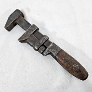 May include: A vintage adjustable wrench featuring a dark, weathered metal head and a textured, oxidized steel body with a knurled adjustment nut. The handle is constructed from dark, worn wood and is secured with a single metal rivet at the base.