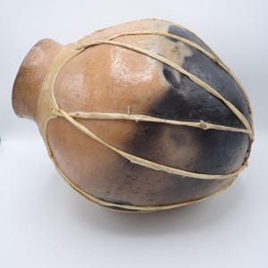 May include: A rounded, bulbous vessel with a smooth, tan and charcoal-black gradient finish. The exterior is wrapped in a decorative network of thin, light-colored, fibrous strips that crisscross over the surface of the container.