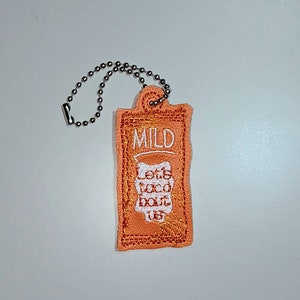 Puede incluir: Un peque&ntilde;o llavero naranja con una cadena plateada. El llavero tiene las palabras "Mild" y "Let's Taco Bout Us" bordadas.
