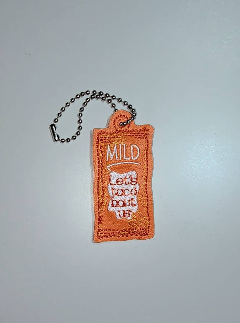 Puede incluir: Un peque&ntilde;o llavero naranja con una cadena plateada. El llavero tiene las palabras "Mild" y "Let's Taco Bout Us" bordadas.