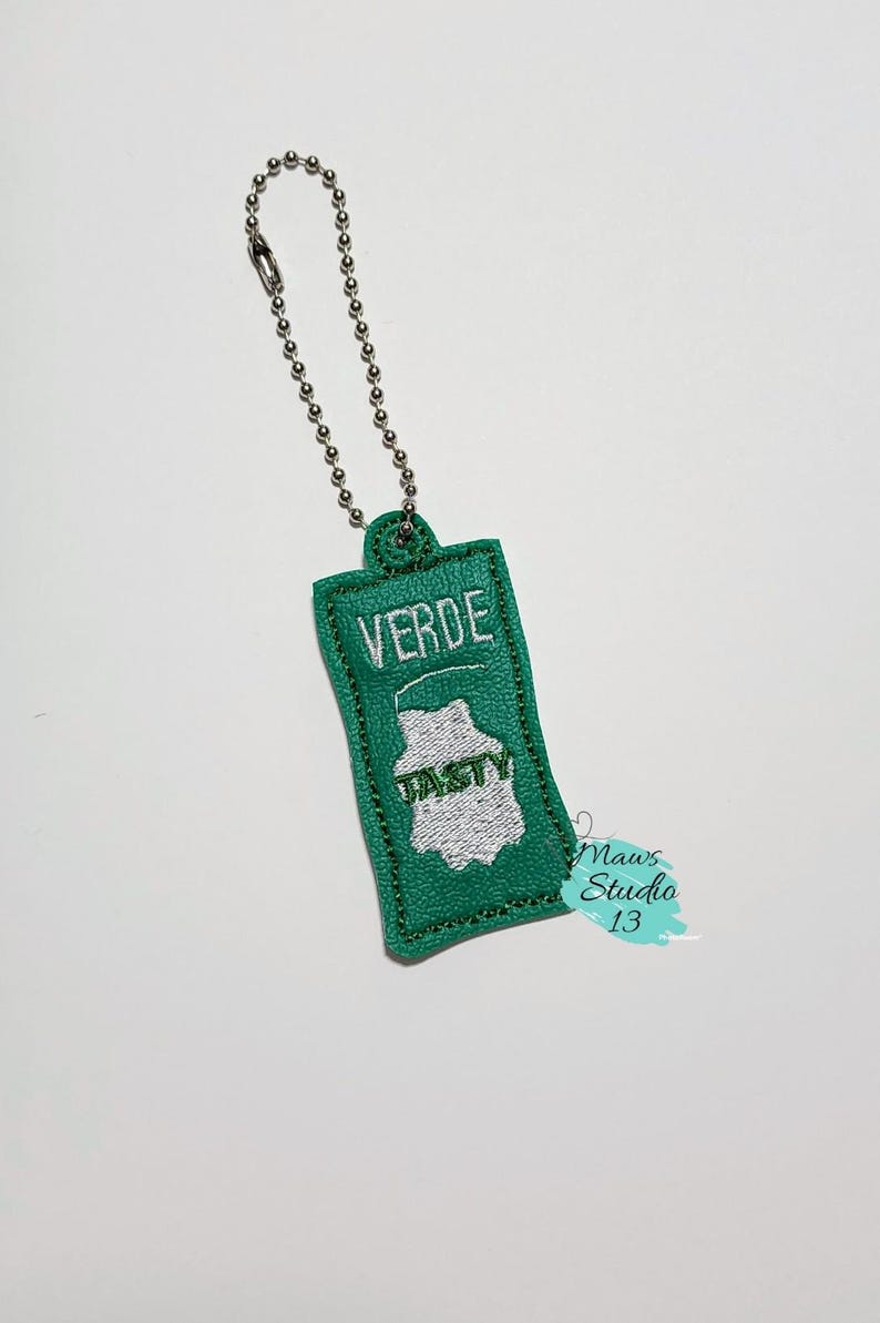 Puede incluir: Un llavero de tela verde con un dise&ntilde;o bordado en blanco que dice "VERDE" y "TASTY". El llavero tiene una cadena plateada.