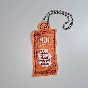 Puede incluir: Llavero de tela naranja con un borde rojo bordado y las palabras "HOT" y "You had me at tacos" en bordado blanco.