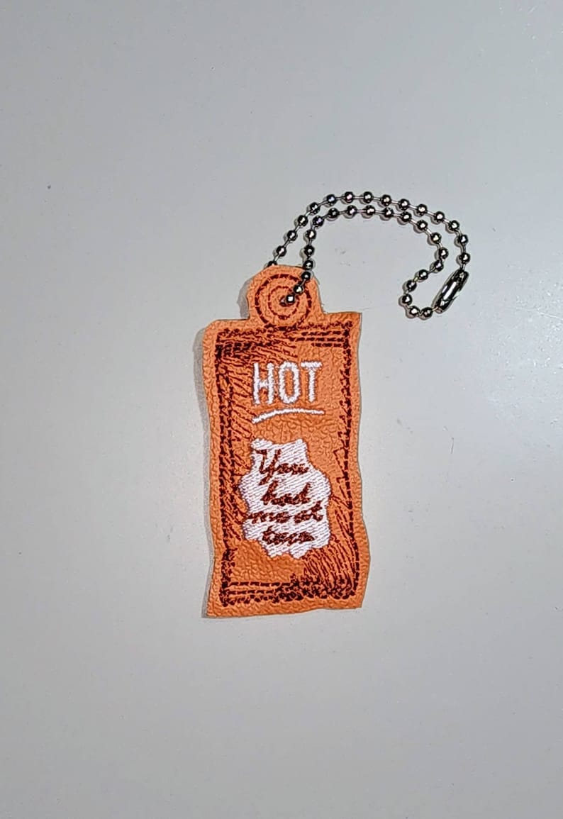 Puede incluir: Llavero de tela naranja con un borde rojo bordado y las palabras "HOT" y "You had me at tacos" en bordado blanco.