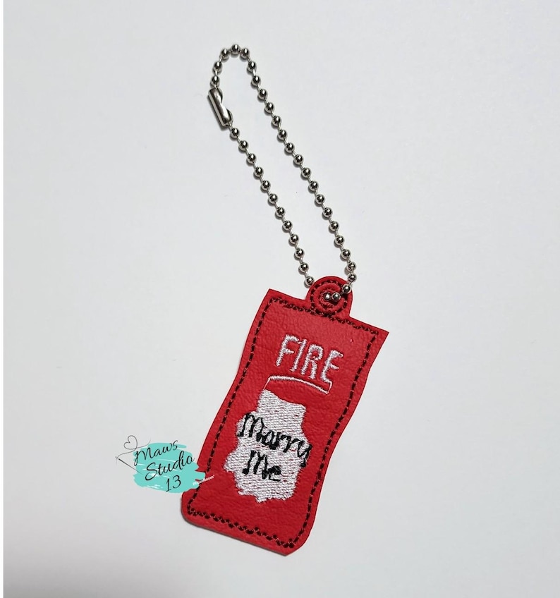 Puede incluir: Llavero de tela roja con forma de botella de salsa picante con las palabras "FIRE" y "Marry Me" bordadas.