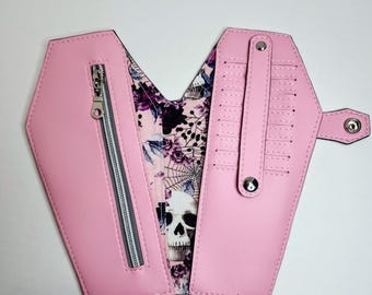 May include: A pink faux leather wallet shaped like a coffin with a skull and spiderweb print lining. The wallet has a zippered pocket and a snap closure.