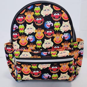 May include: Black backpack featuring a repeating pattern of Muppet characters. The backpack has a black top and trim, a front zippered pocket, and side pockets. The design includes Kermit the Frog, Miss Piggy, and Fozzie Bear.