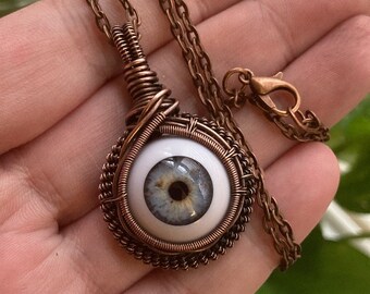 May include: A copper wire wrapped pendant necklace featuring a blue glass eye with a black pupil. The necklace has a copper chain with a lobster clasp.