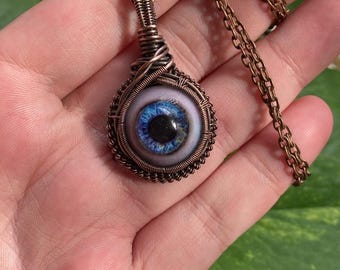 May include: A copper wire wrapped pendant necklace featuring a blue eye with a white sclera and a black pupil. The necklace is on a copper chain.