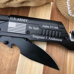 May include: A black multi-tool knife with the text "U.S. ARMY", an American flag, and the words "Be Safe". The knife also has the text "Alpha 3/116th" and "Sergeant J. Anderson". The knife is on a wooden surface.