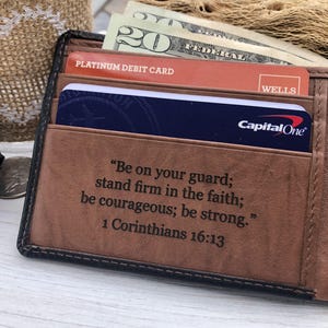 May include: A brown leather bifold wallet featuring an engraved quote from 1 Corinthians 16:13. The wallet contains a Capital One credit card, a Wells Fargo platinum debit card, and a twenty-dollar bill tucked into the interior card slots.