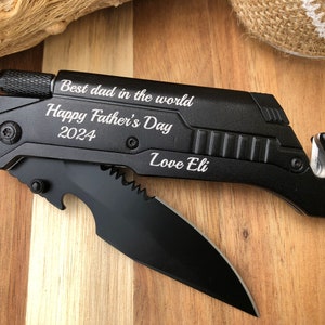 May include: Black multi-tool with a knife blade, bottle opener, and other tools. The text "Best dad in the world, Happy Father's Day 2024, Love Eli" is engraved on the tool. The tool is on a wooden surface.