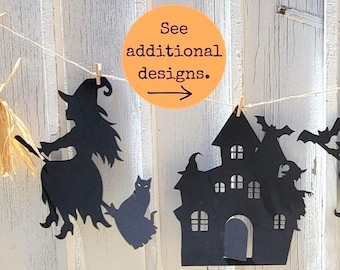 May include: Black paper Halloween garland featuring witch silhouettes, a haunted house, and a cat. The garland is strung on twine and held with small wooden clothespins. An orange circle with the text "See additional designs" is also included.