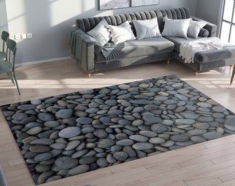 May include: A rectangular rug with a pattern of smooth, grey river stones. The rug is placed on a light-coloured hardwood floor, with a grey sectional sofa and a dining table visible in the background. The rug adds a natural, textured element to the room.