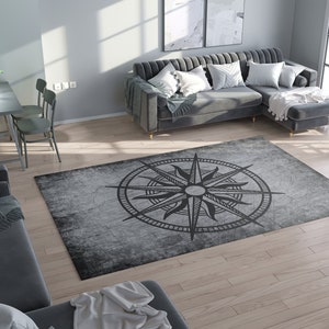 May include: A rectangular gray area rug with a compass rose design in black. The rug has a distressed, vintage look, with a faded appearance and a dark border. The compass rose features directional points and a central starburst.