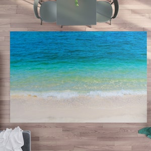 May include: A rectangular rug featuring a beach scene with turquoise water and a white sandy shore. The rug has a top-down perspective, with a wave pattern at the bottom. The rug is placed on a wooden floor, with furniture visible in the background.
