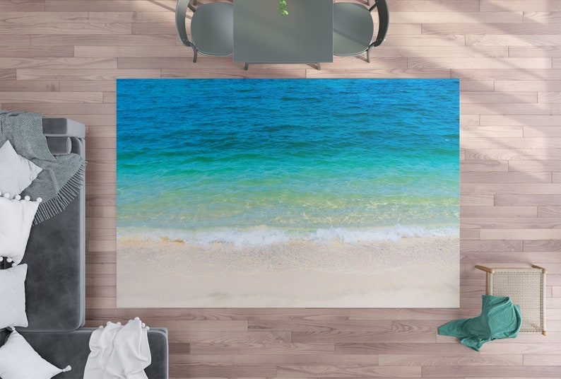 May include: A rectangular rug featuring a beach scene with turquoise water and a white sandy shore. The rug has a top-down perspective, with a wave pattern at the bottom. The rug is placed on a wooden floor, with furniture visible in the background.