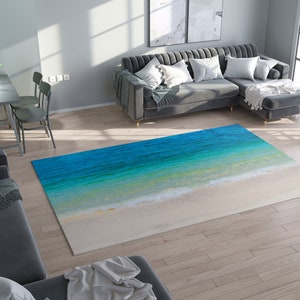 May include: A rectangular area rug featuring a beach scene with turquoise water and white sand. The rug's design mimics the ocean's edge, with varying shades of blue and green transitioning to a sandy white at the bottom. The rug is placed on a hardwood floor.